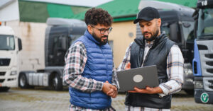 two people having a discussion and looking at computer next to a few semi trucks