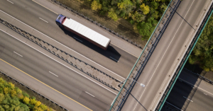 Aerial view of a semi-truck traveling on a multi-lane highway, passing under a concrete overpass surrounded by green and yellow trees.